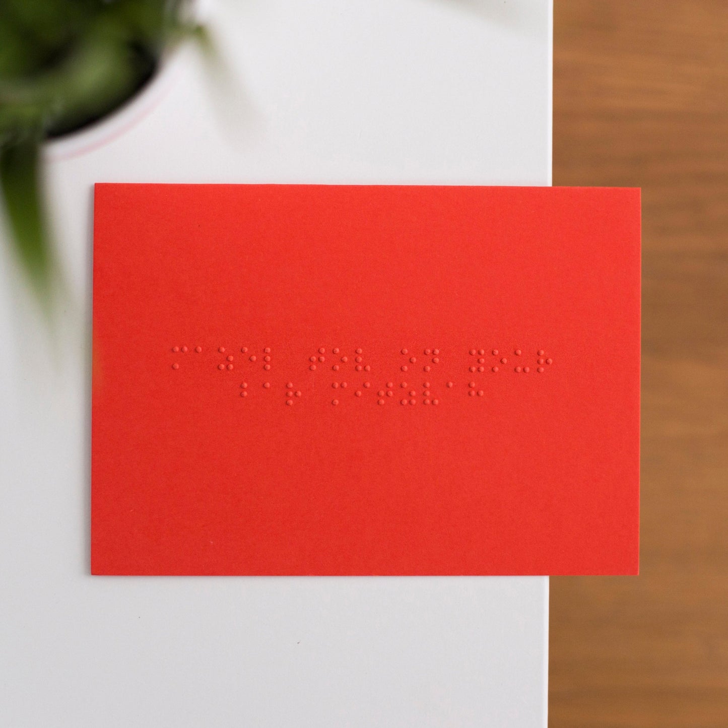 A vibrant red card lay flat on a table with mazel tov on your bar mitzvah written in lower case grade one braille. 