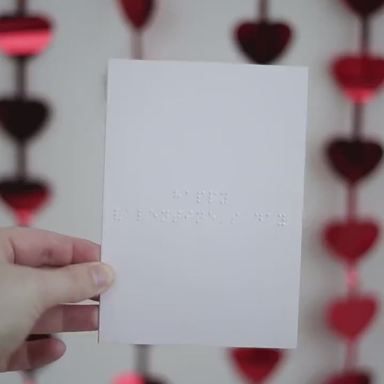 A video of a pale pink card with Happy Valentine's Day written in braille. There are hanging red shiny love hearts in the background.