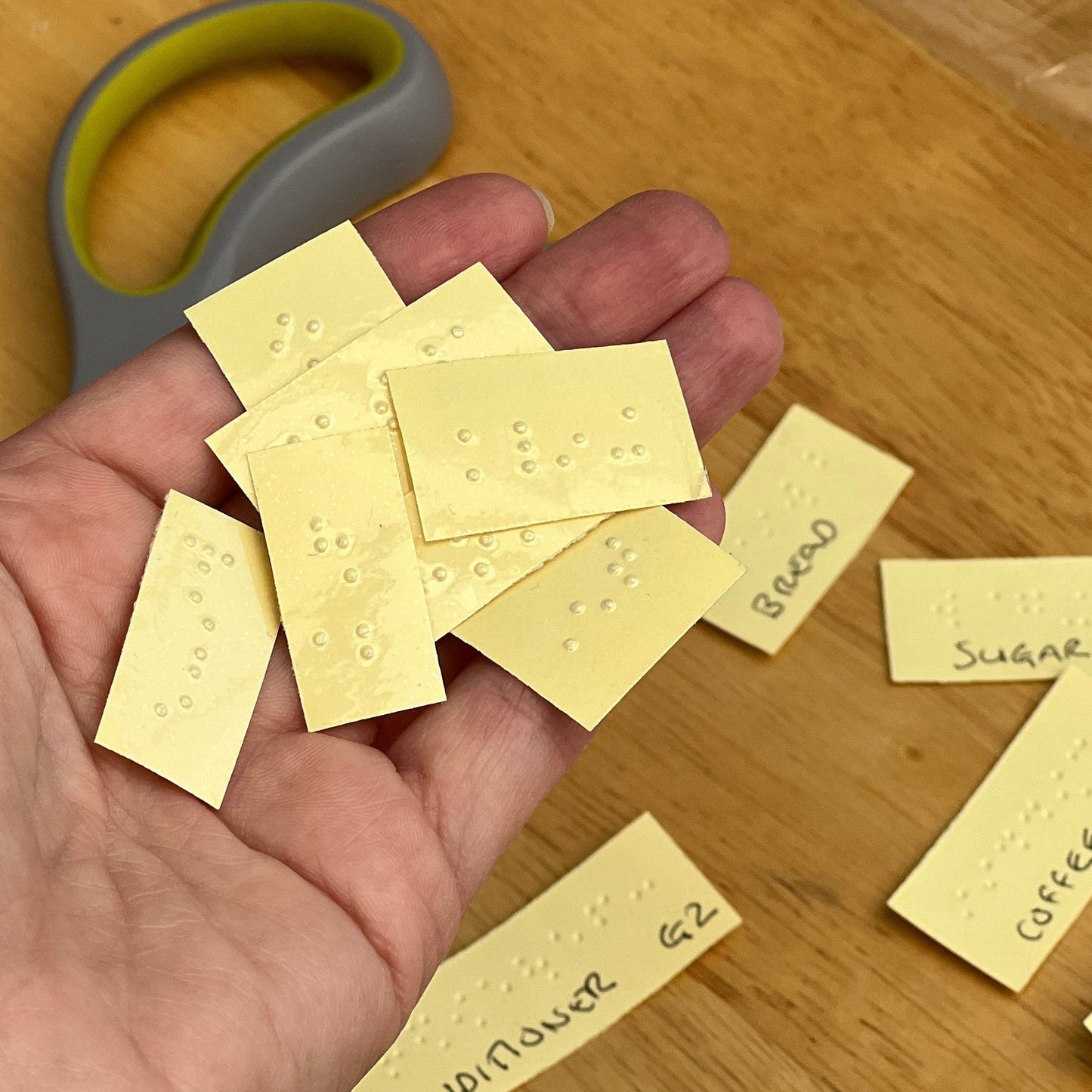 Image shows my hand holding some of the braille labels I have printed, in the background you can see the reverse of the labels where I have written what each labels says and whether grade 1 or grade 2. You can also see the top of a pair of scissors.