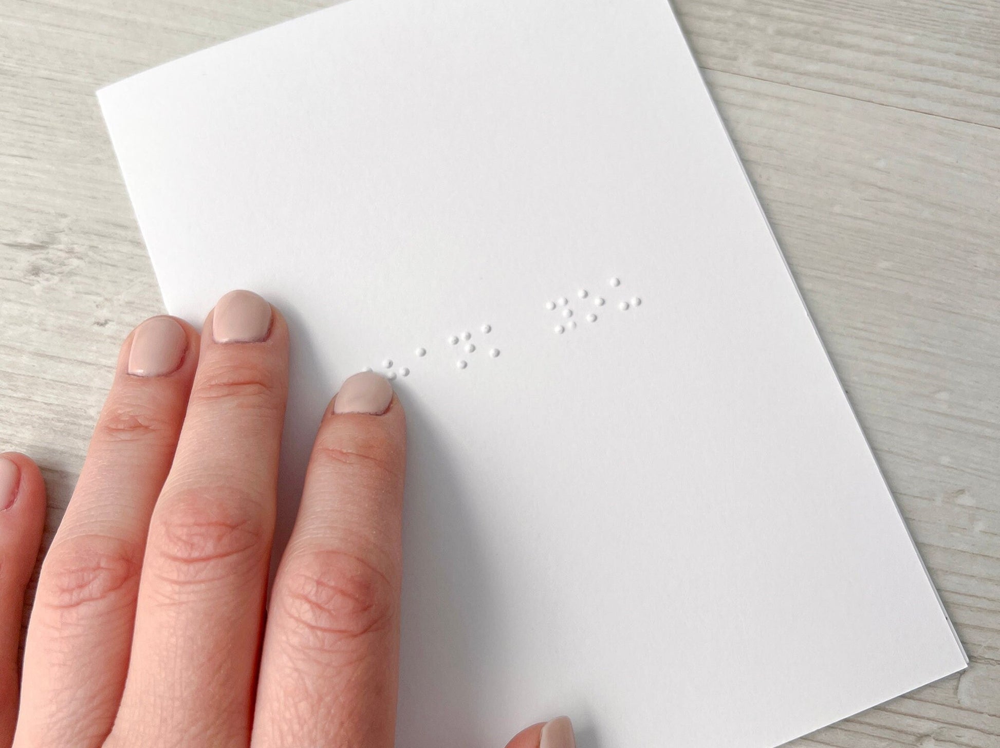 A white braille card with thank you written in lower case braille, a hand wearing pink nail varnish is on top of the card