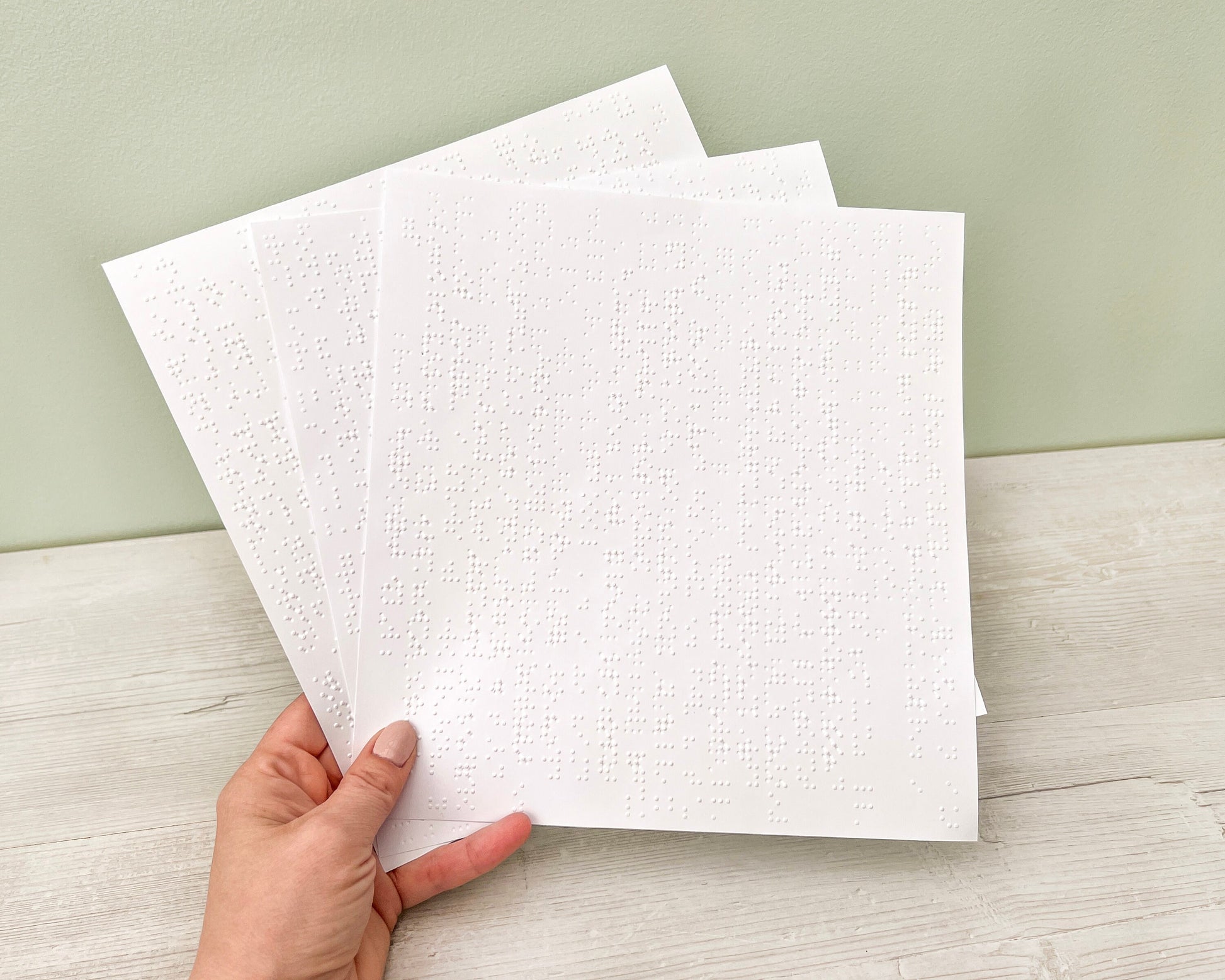 Three sheets of 22x23cm white braille paper are being held by a hand.