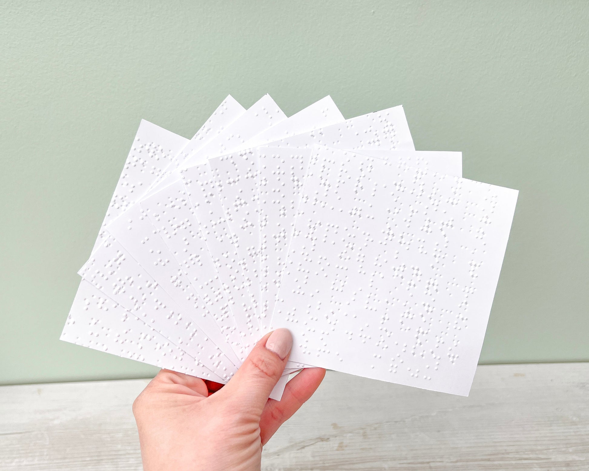 A hand holding 8 sheets of braille paper, approx. 11 x 11cm each.
