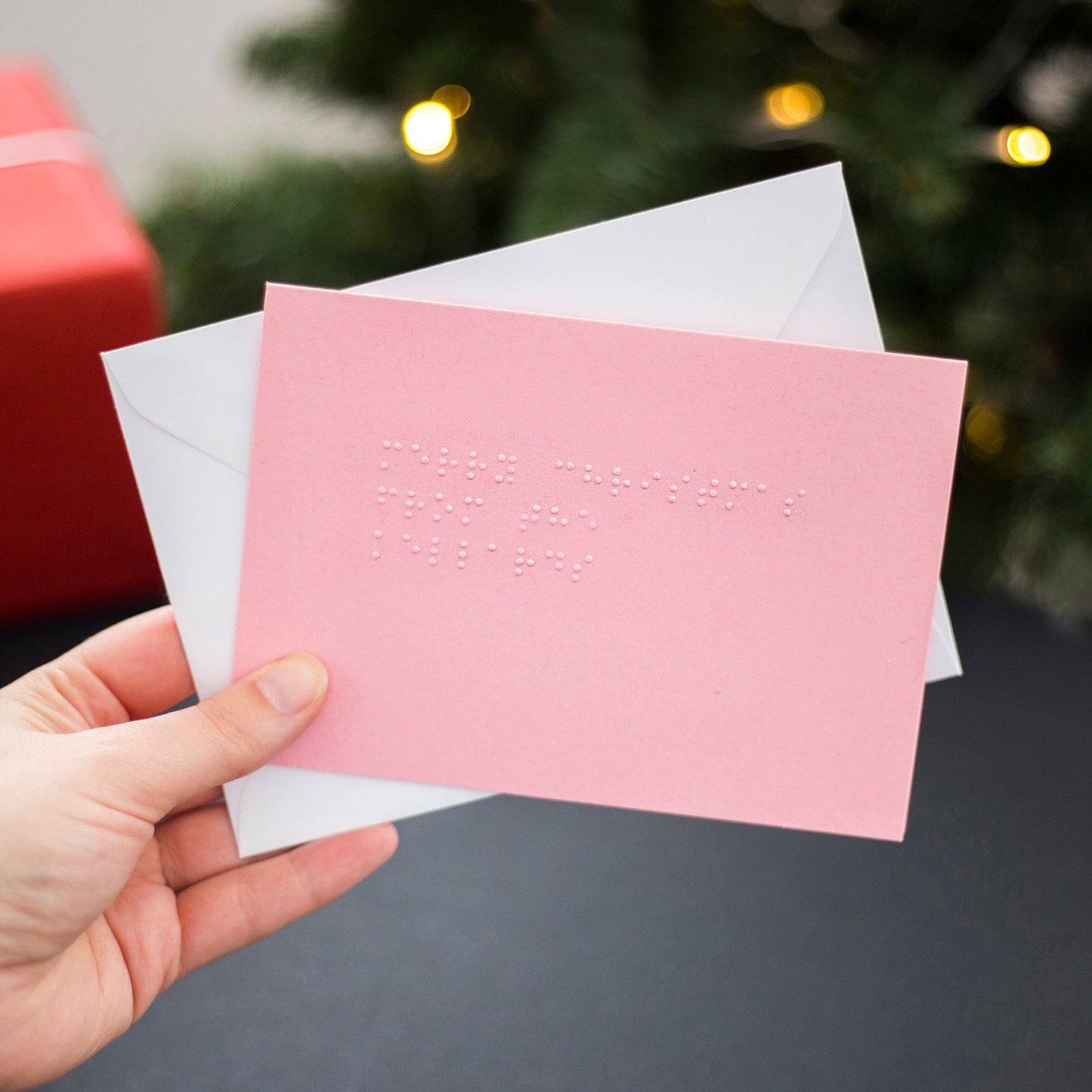A hand holding a pink braille Christmas card with Merry Christmas from the Kellards written in lower case UEB.