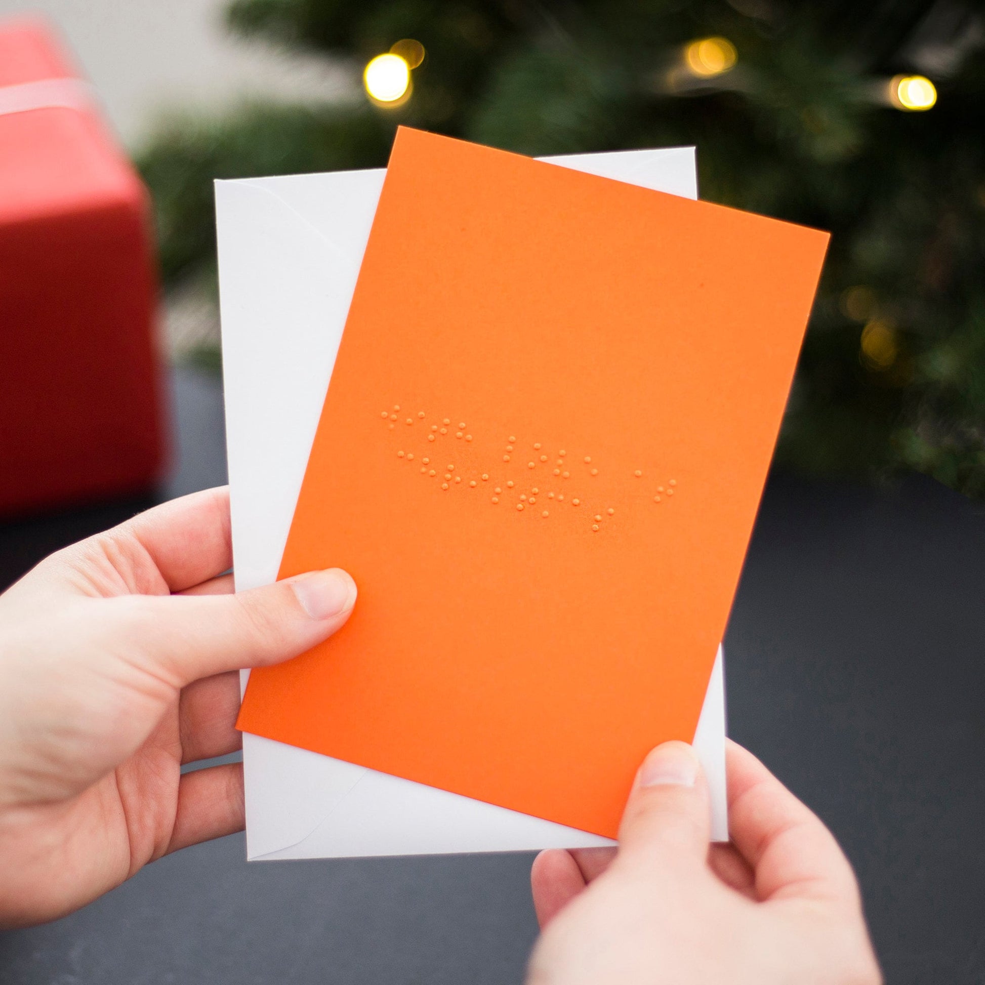 Hands holding an orange braille Christmas card saying With Love at Christmas written in lower case UEB.