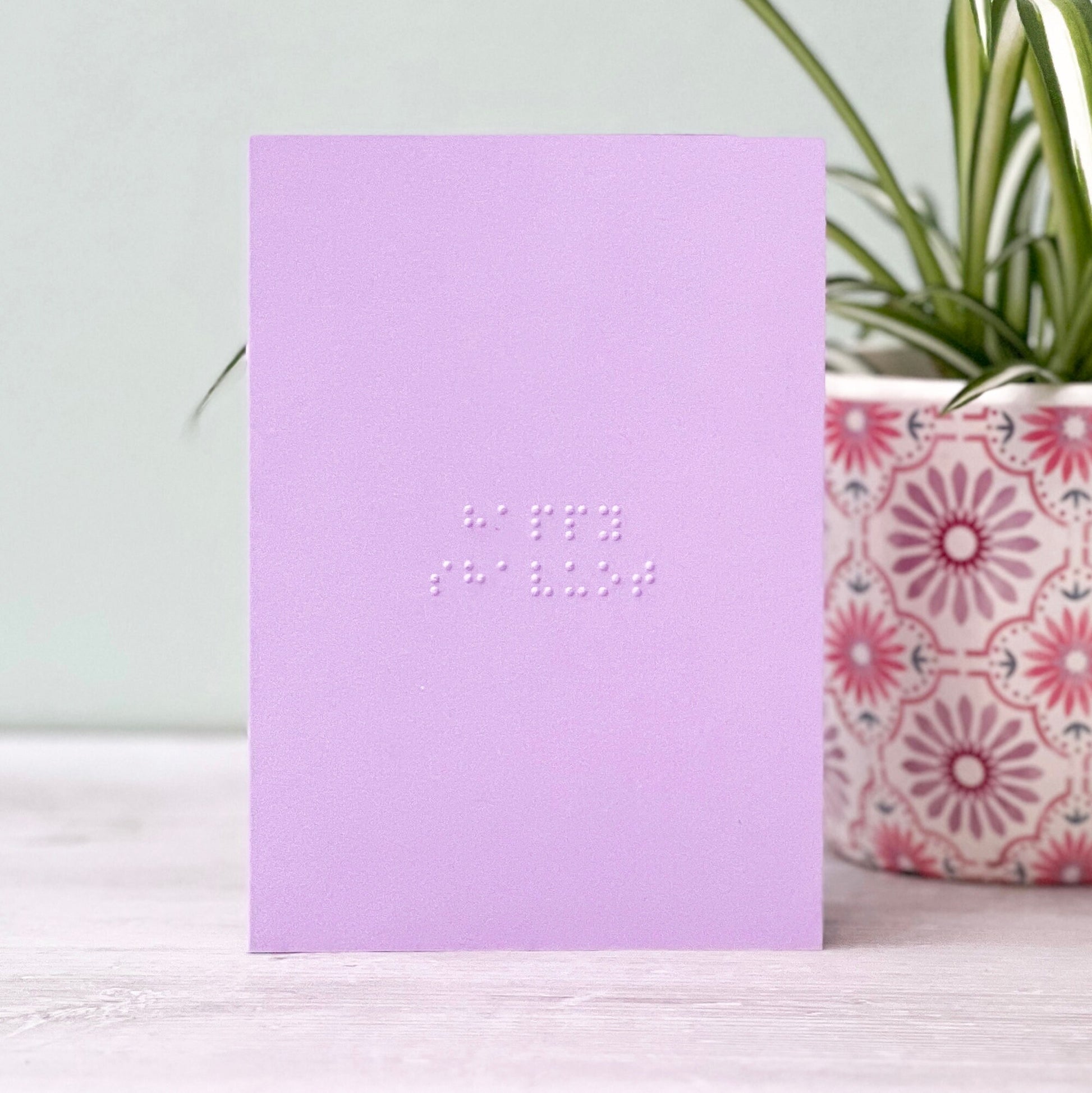 A pastel purple greetings card with happy shavuot written in grade 1 braille. There is a plant to the right of the photo.