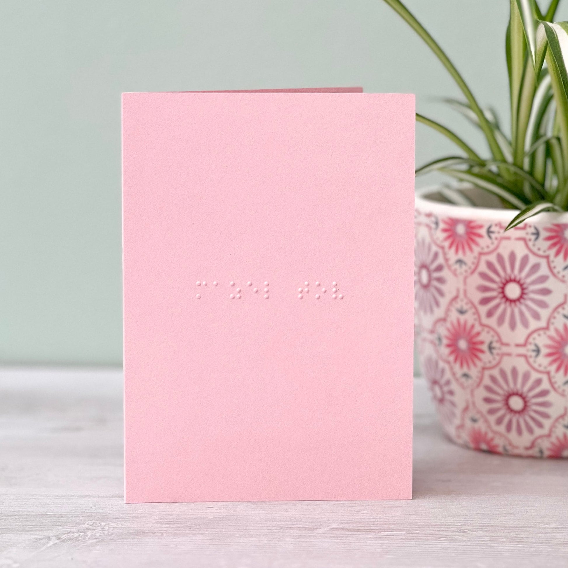 A pastel pink greetings card with mazel tov written in grade 1 braille. There is a plant to the right of the photo.