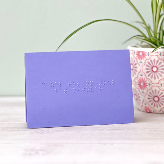 A vibrant purple greetings card with mazel tov on your bat mitzvah written in grade 1 braille. There is a plant to the right of the photo.