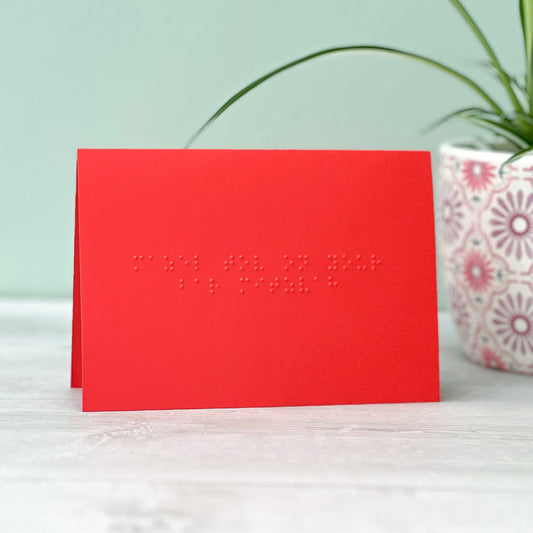 A vibrant red greetings card with mazel tov on your bar mitzvah written in grade 1 braille. There is a plant to the right of the photo.