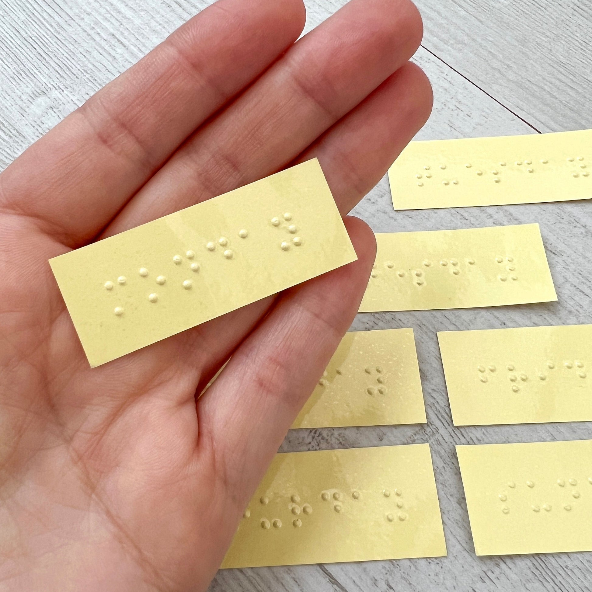 A hand holding a braille label which says monday, in the background are other braille labels with the days of the week on.