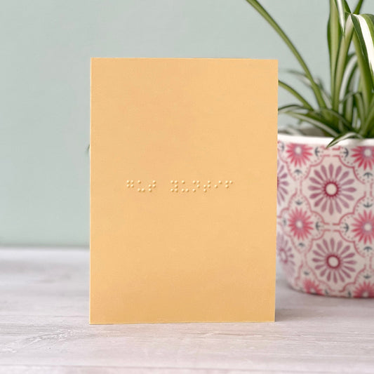 A pastel yellow greetings card with gut yuntif written in grade 1 braille. There is a plant to the right of the photo.
