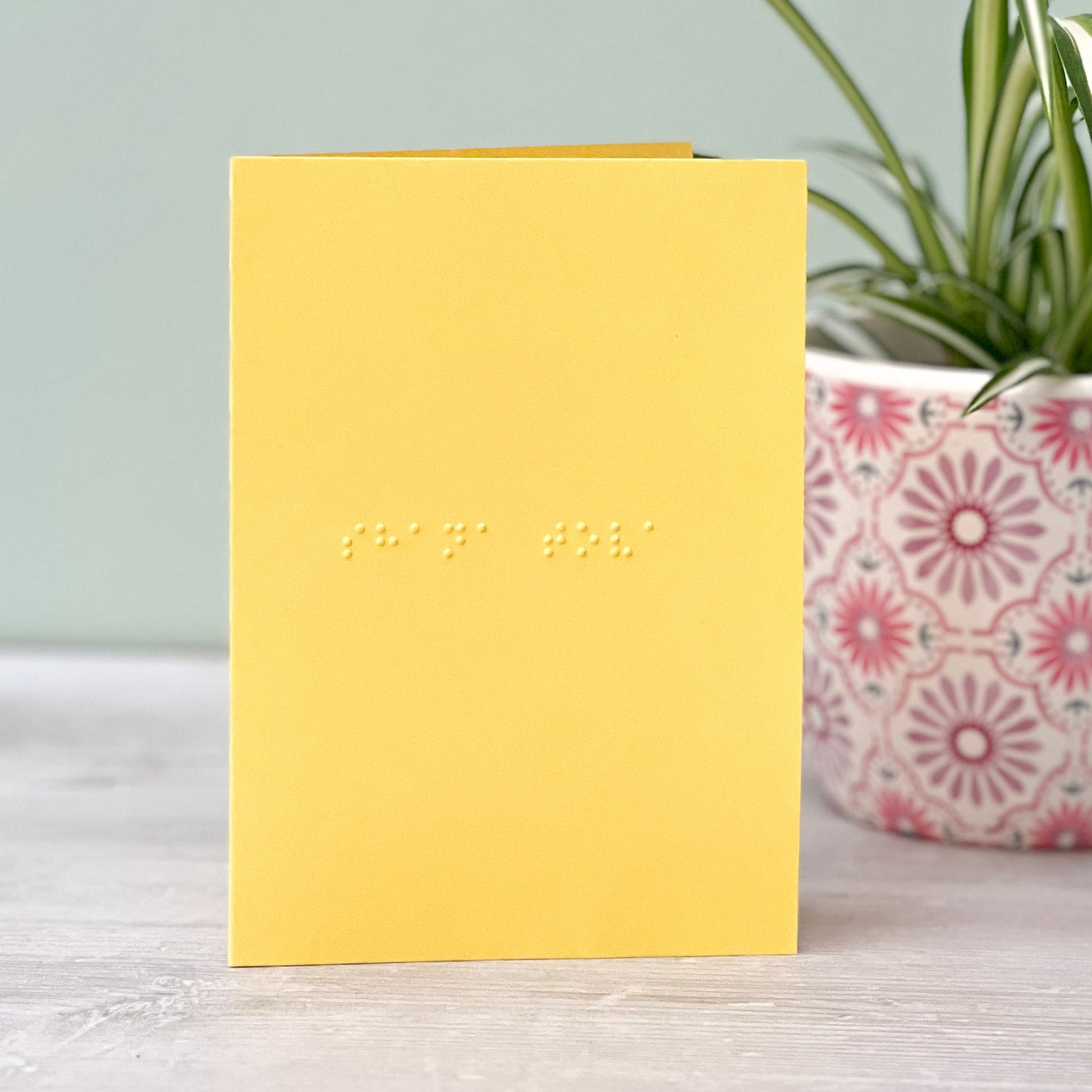 A vibrant yellow greetings card with shana tova written in grade 1 braille. There is a plant to the right of the photo.