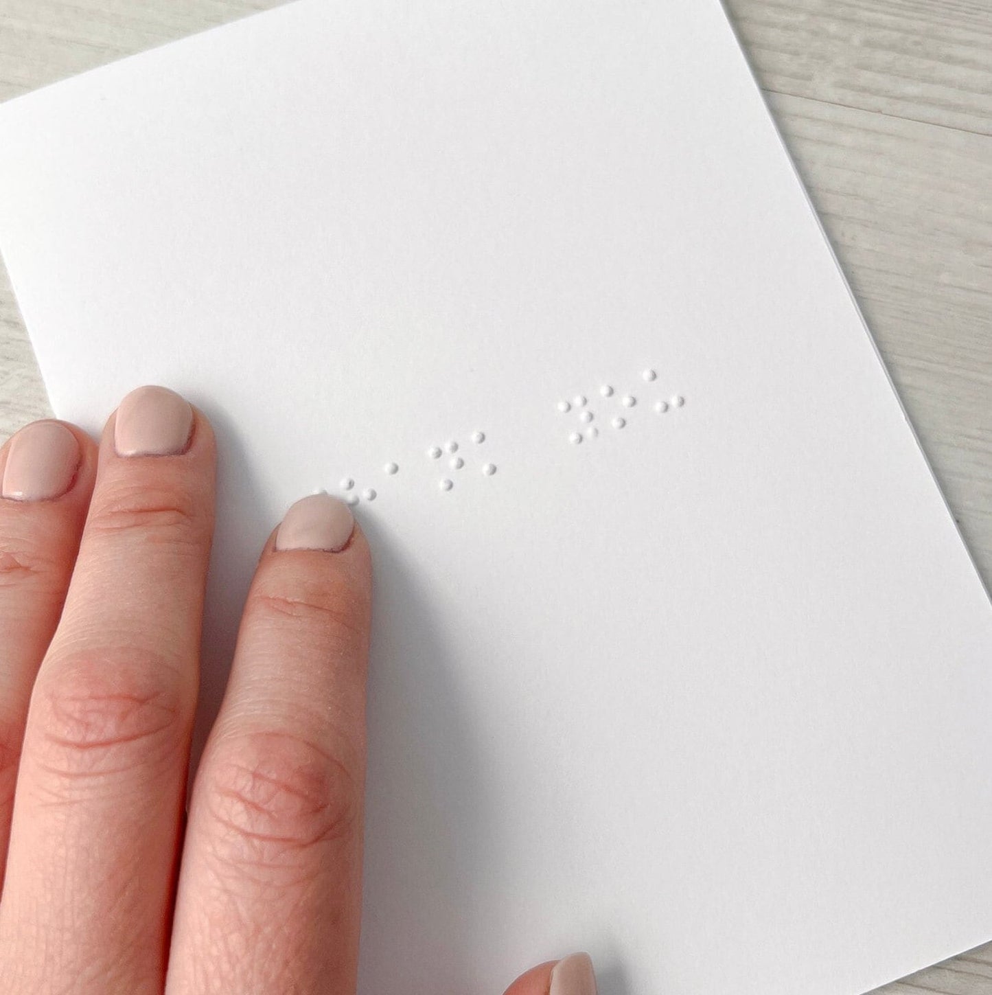 A white braille card with thank you written in lower case braille, a hand wearing pink nail varnish is on top of the card